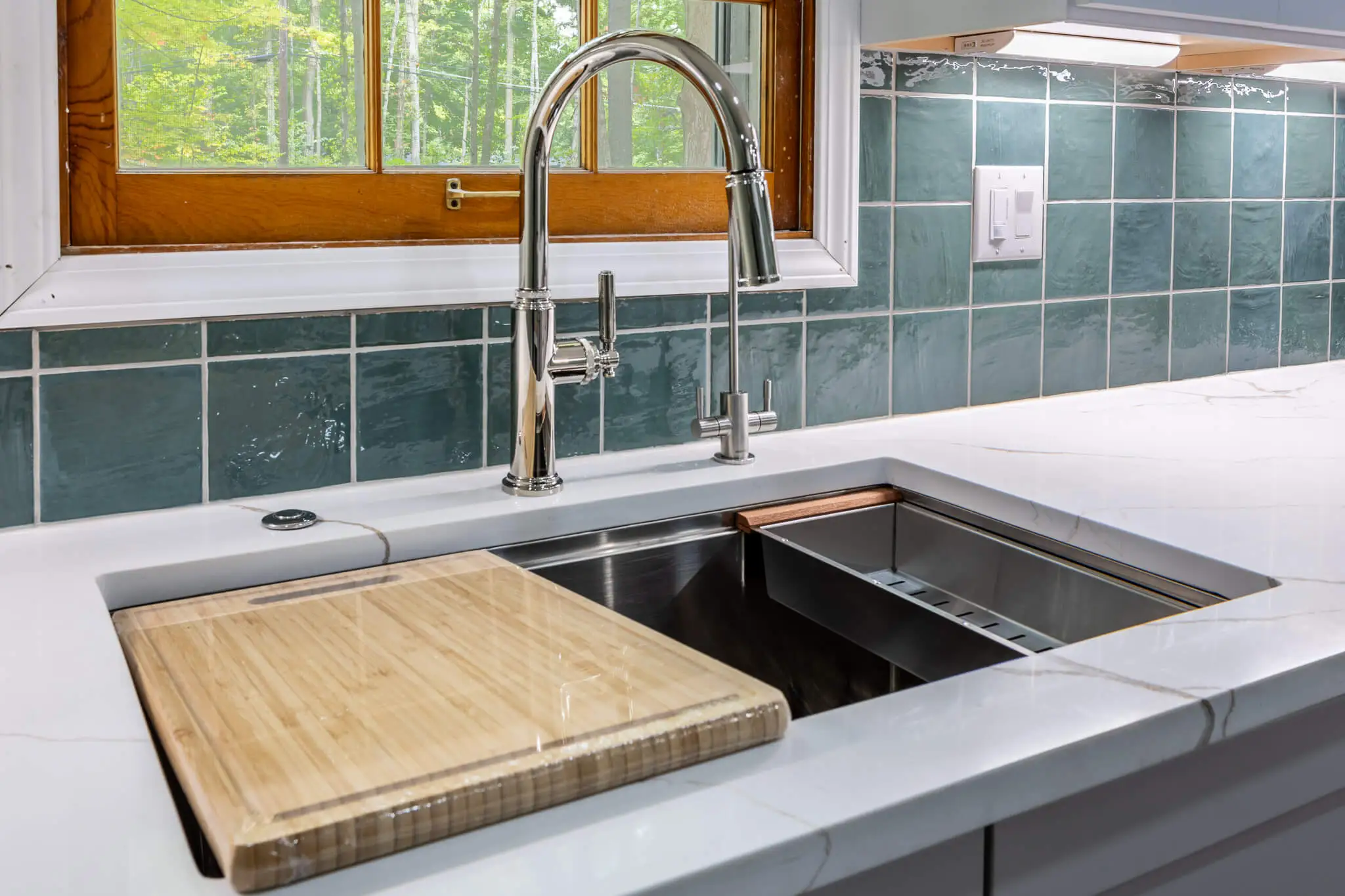 Close-up of a stainless steel kitchen sink and modern chrome faucet with green tile backsplash and quartz countertop.